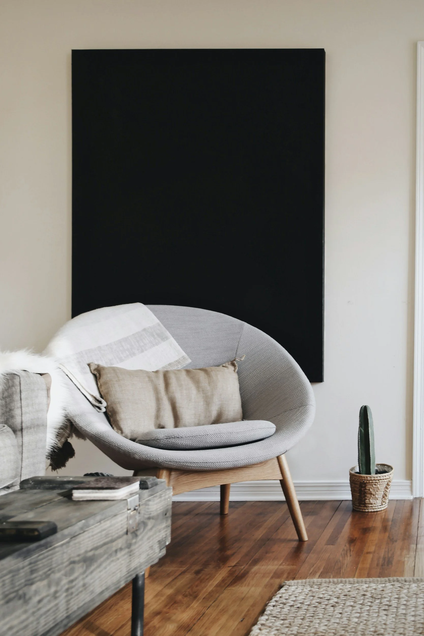 Living room corner with a gray curved lounge chair, beige cushions, striped throw blanket, tall cactus in a woven basket, wooden floor, black wall art, and part of a wooden coffee table.
