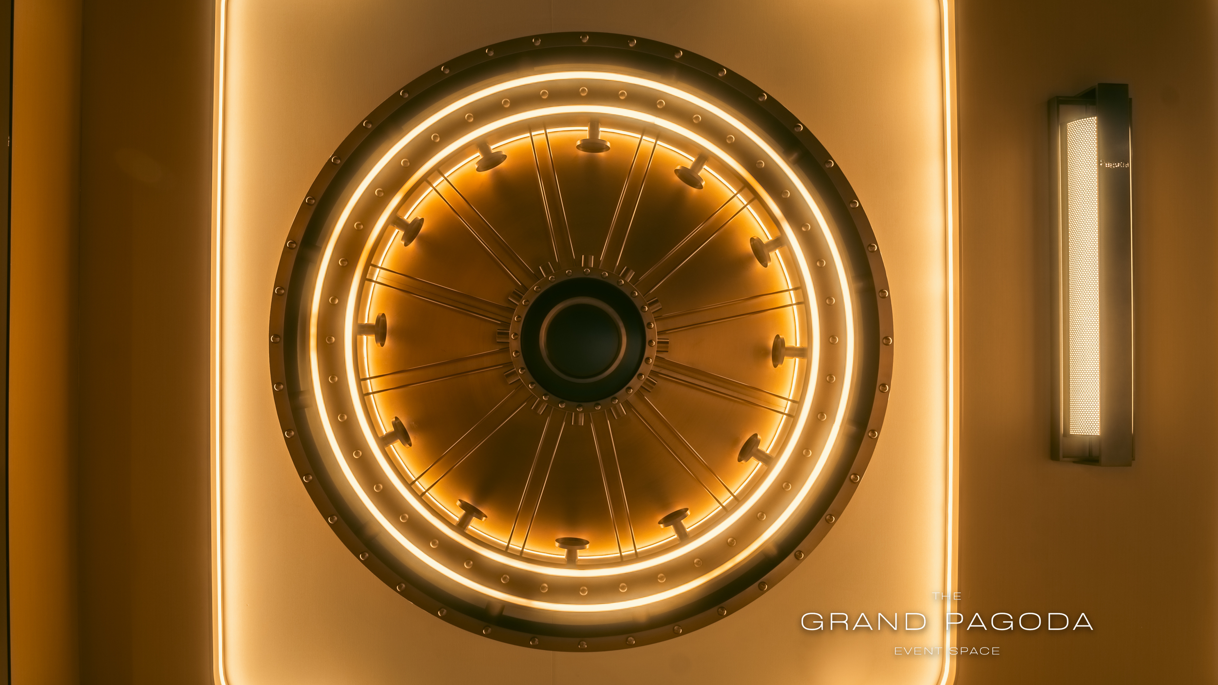 Lighting fixture with circular design and a warm glow, with text overlay reading 'The Grand Pagoda Event Space' on a modern beige wall.