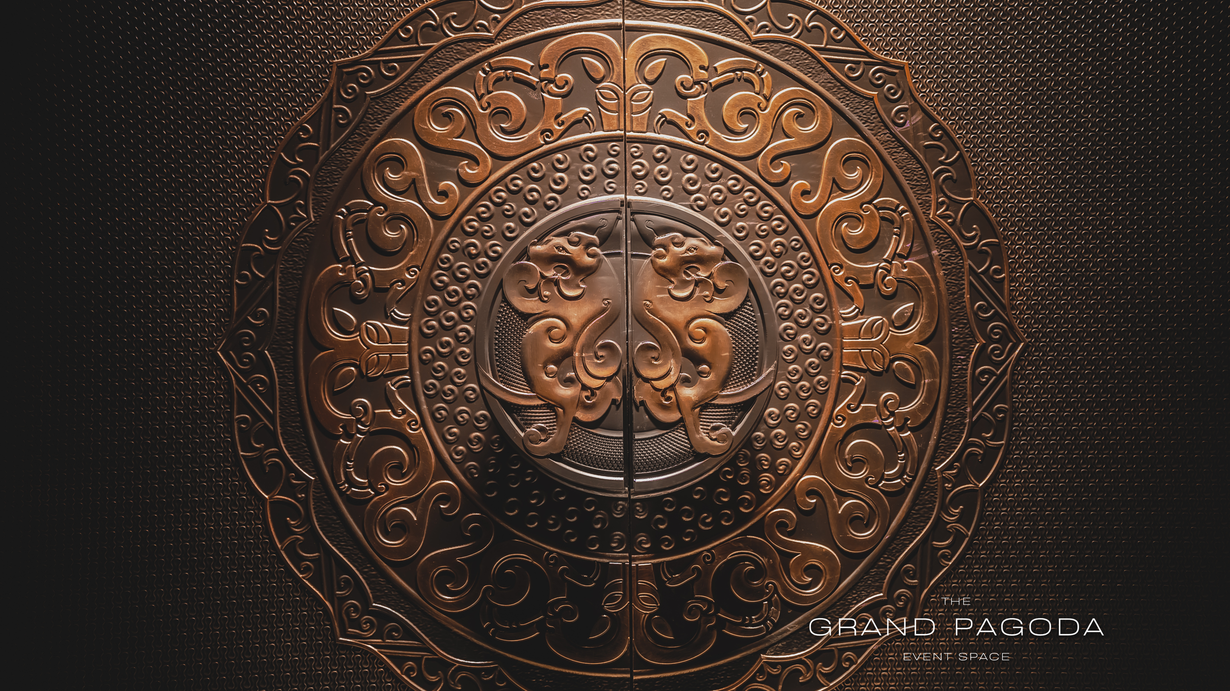 Intricate bronze-colored decorative door with symmetrical swirling patterns and two lion or dragon figures facing each other in the center. Text at the bottom right reads 'The Grand Pagoda Event Space'.