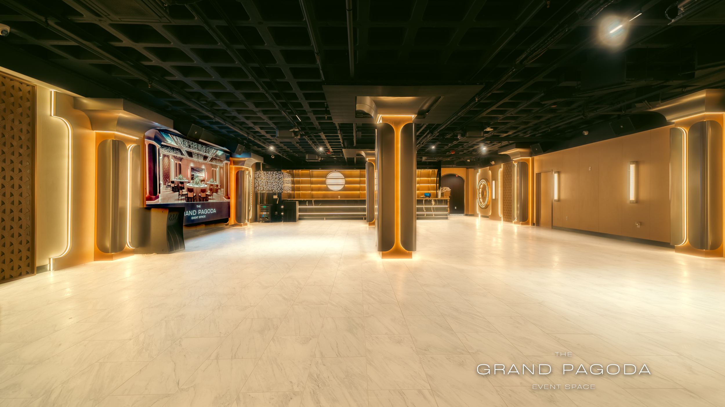 Empty event space with modern decor, illuminated by warm lighting, featuring a large screen, bar area, and open floor with marble flooring.