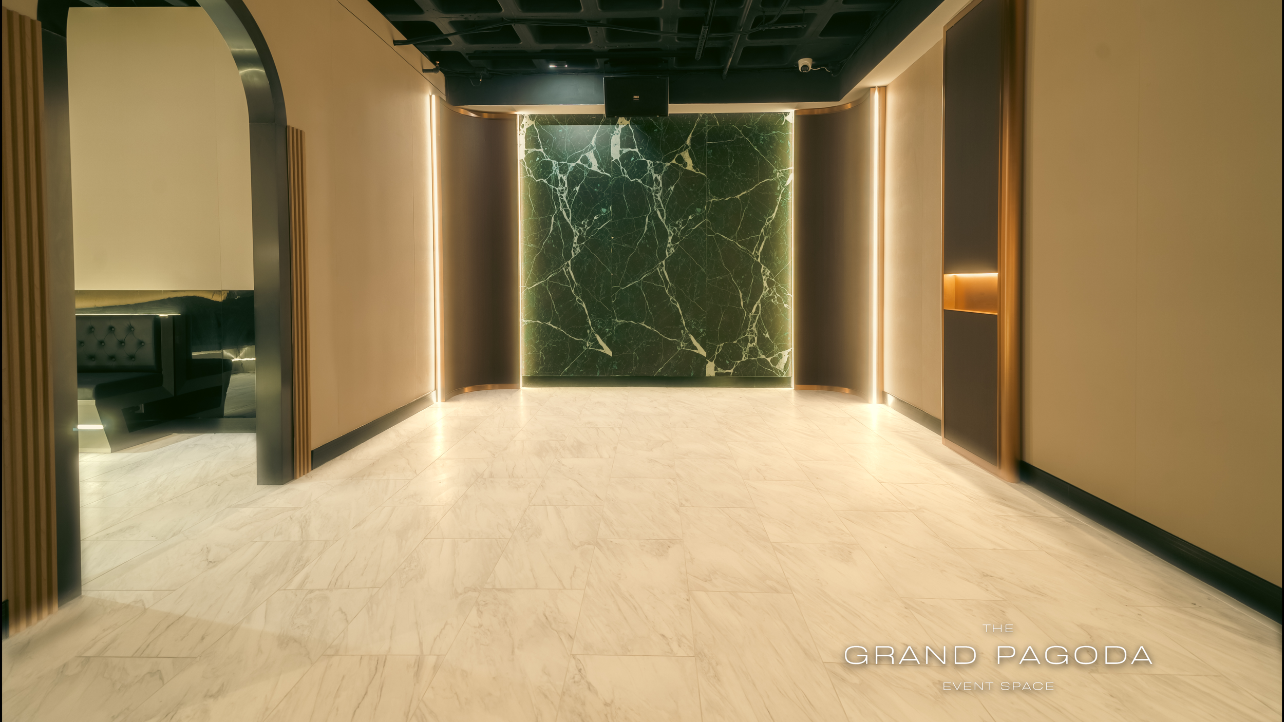 Interior of a modern event space with a large green marble wall, framed with vertical strip lighting, and beige walls with wooden accents. The floor is made of light-colored marble tiles, and there is a black ceiling with some visible lighting and a 