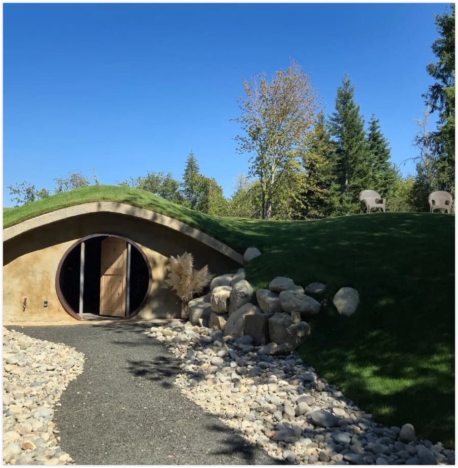 A nature scene featuring a Hobbit-style underground house with a rounded door, surrounded by trees, rocks, and grassy hills under a clear blue sky.
