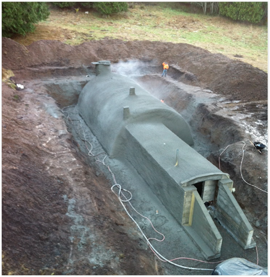 A large underground concrete structure resembling part of a tunnel or storage tank being constructed in a dug-out area with a worker standing nearby.