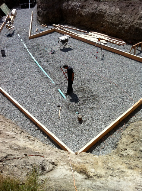Construction site with a person working on the foundation, using a level on a blue pipe, surrounded by wooden frames and construction tools.