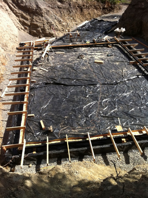 Construction site with wooden formwork and black plastic sheeting in a dug-out area for foundation work.