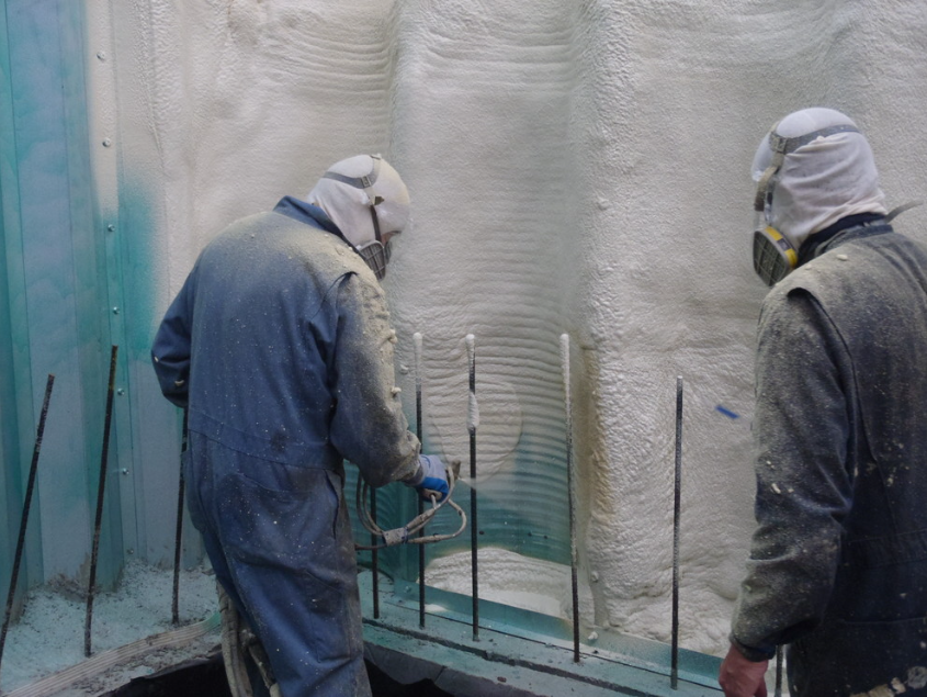 Two workers in protective gear and dust masks spray foam insulation onto a wall under construction.