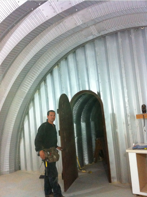 A man in a workshop standing inside a large, metallic, arched structure with a wooden door open and leading into a smaller, tunnel-like space.