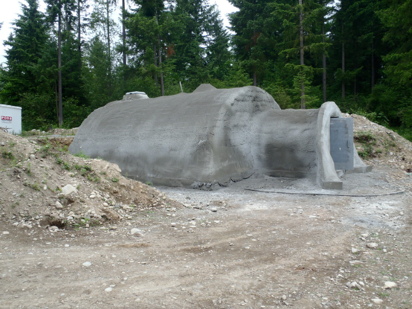 A large, unpainted concrete structure that resembles a tunnel or bunker under construction, with a forested area in the background.