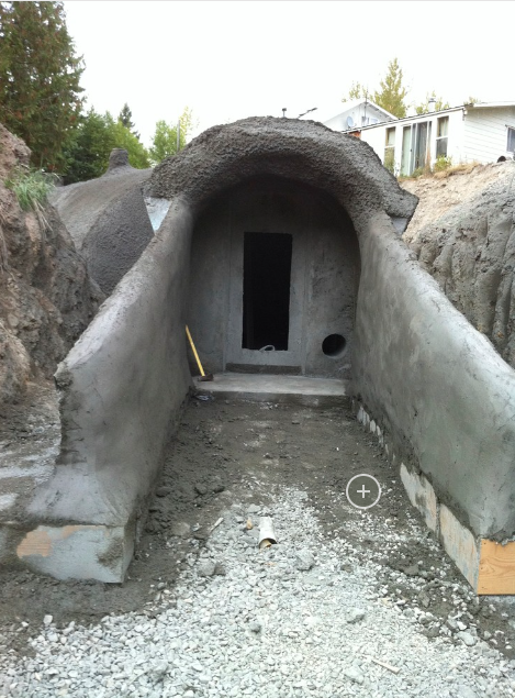 Underground concrete tunnel with a rounded ceiling, leading to a dark doorway, surrounded by construction site dirt and gravel.