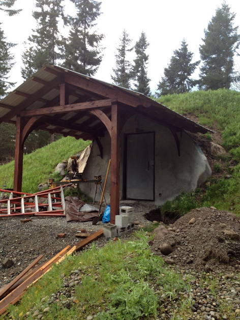 A small shelter with a wooden roof supported by wooden posts, built on a grassy hillside with tall trees in the background. The shelter is made of a concrete structure with a door, and construction materials, including a ladder, are scattered around.