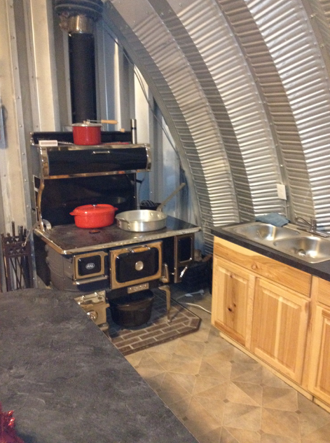Inside a rustic kitchen with a vintage black stove, red and silver pots on top, a wooden cabinet, and corrugated metal walls.