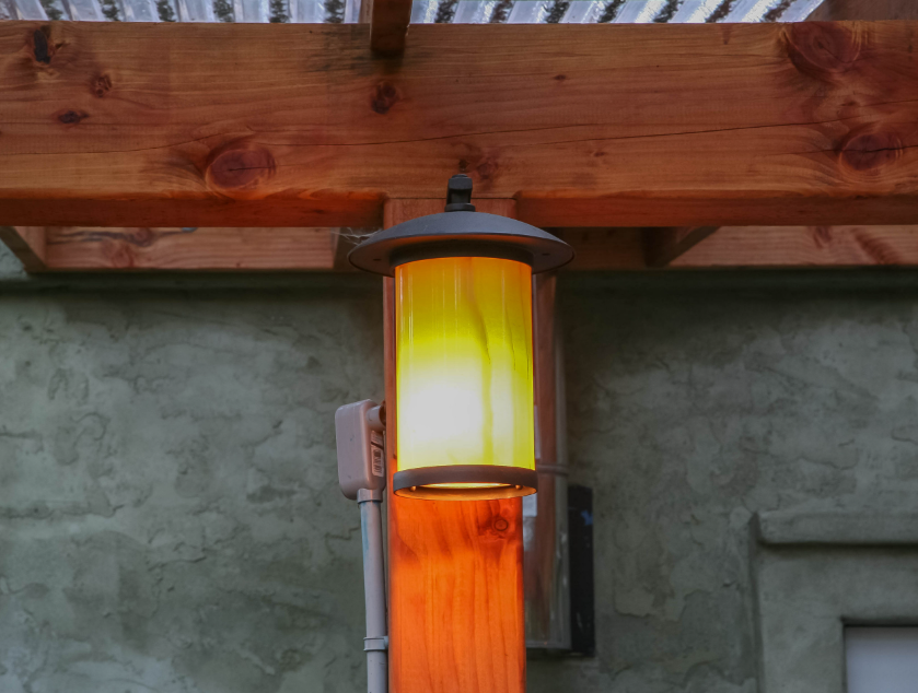 A modern outdoor light fixture mounted on a wooden post, emitting a warm yellow glow, with a wooden deck overhead and a textured stucco wall in the background.
