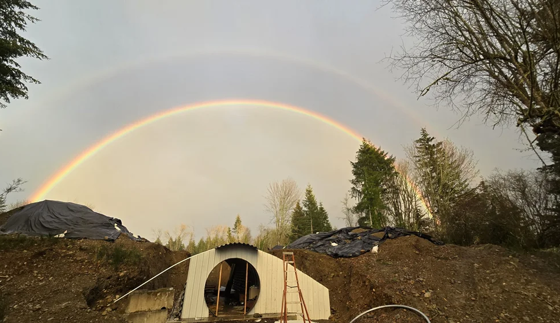 Rainbow over a construction site with a small, partially built structure, trees, and dirt mounds.