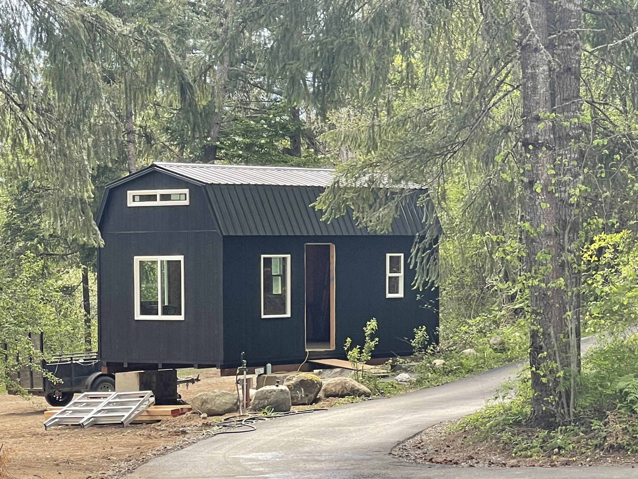 Small black house under construction in a wooded area, with a metal roof, surrounded by trees and rocks, and a paved pathway leading up to it.
