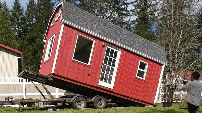 A small red shed with a gray roof tilted on its side on a trailer in a backyard.