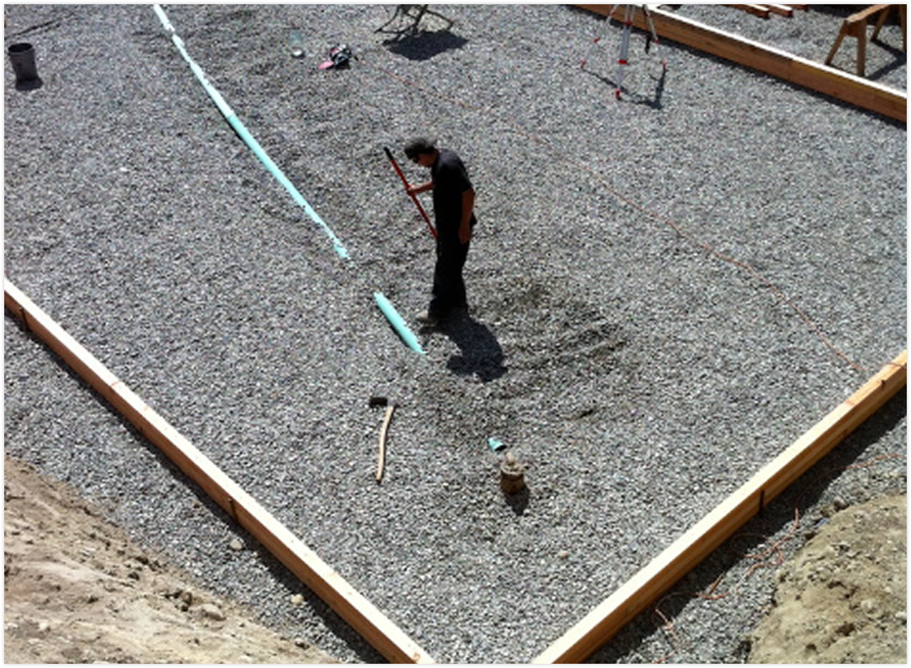 Construction worker standing in a gravel lot, installing a light blue pipe inside a rectangular wooden frame, with various tools and construction materials scattered around.