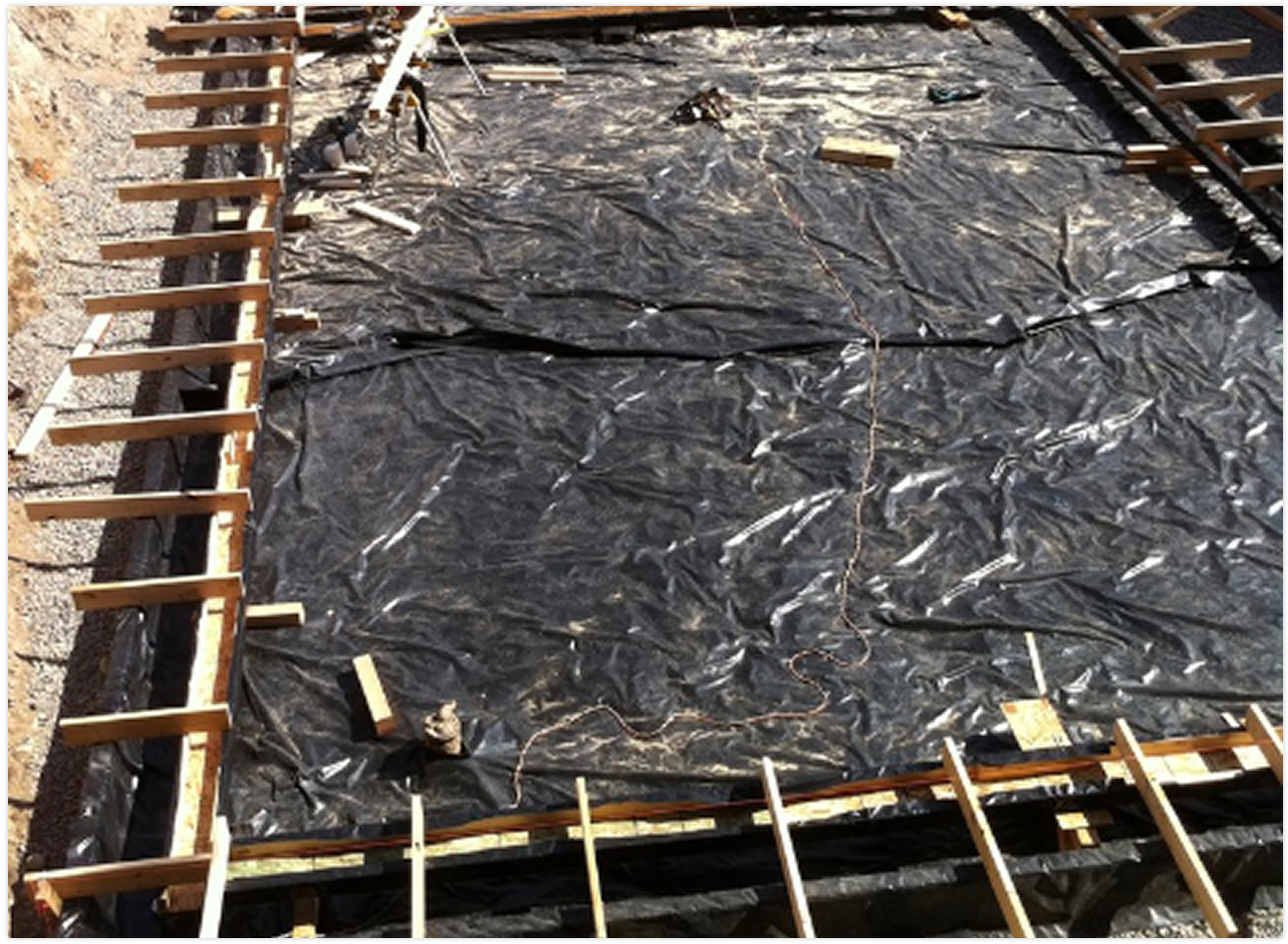 Construction site with black plastic sheeting covering the ground, wooden framework around the perimeter, and scattered construction tools and materials.