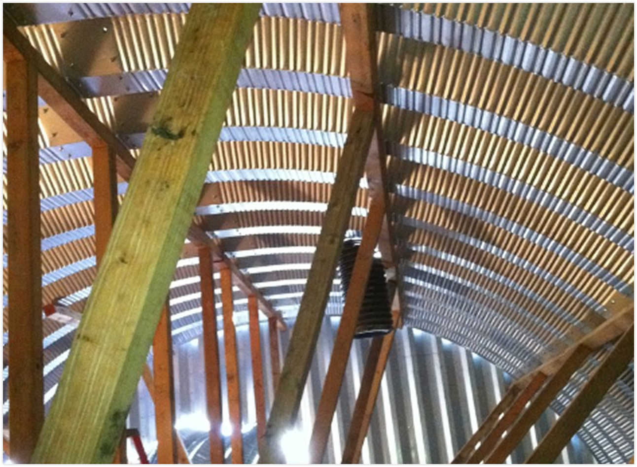 View of the interior of a barn or similar wooden structure, showing unfinished wood framing, curved corrugated metal roofing, and sunlight filtering through gaps.