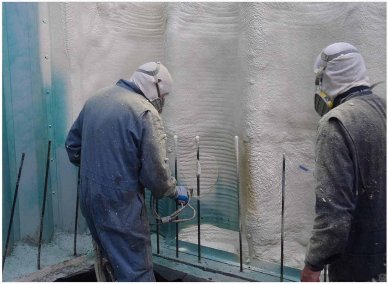 Two workers wearing protective suits and masks spray foam insulation on a wall.