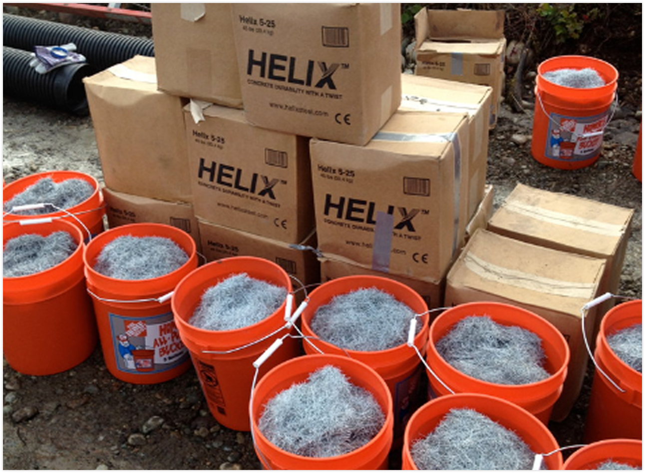 Multiple orange buckets filled with steel wool and several cardboard boxes labeled HELIX stacked on a construction site with dirt ground and black drainage pipes in the background.