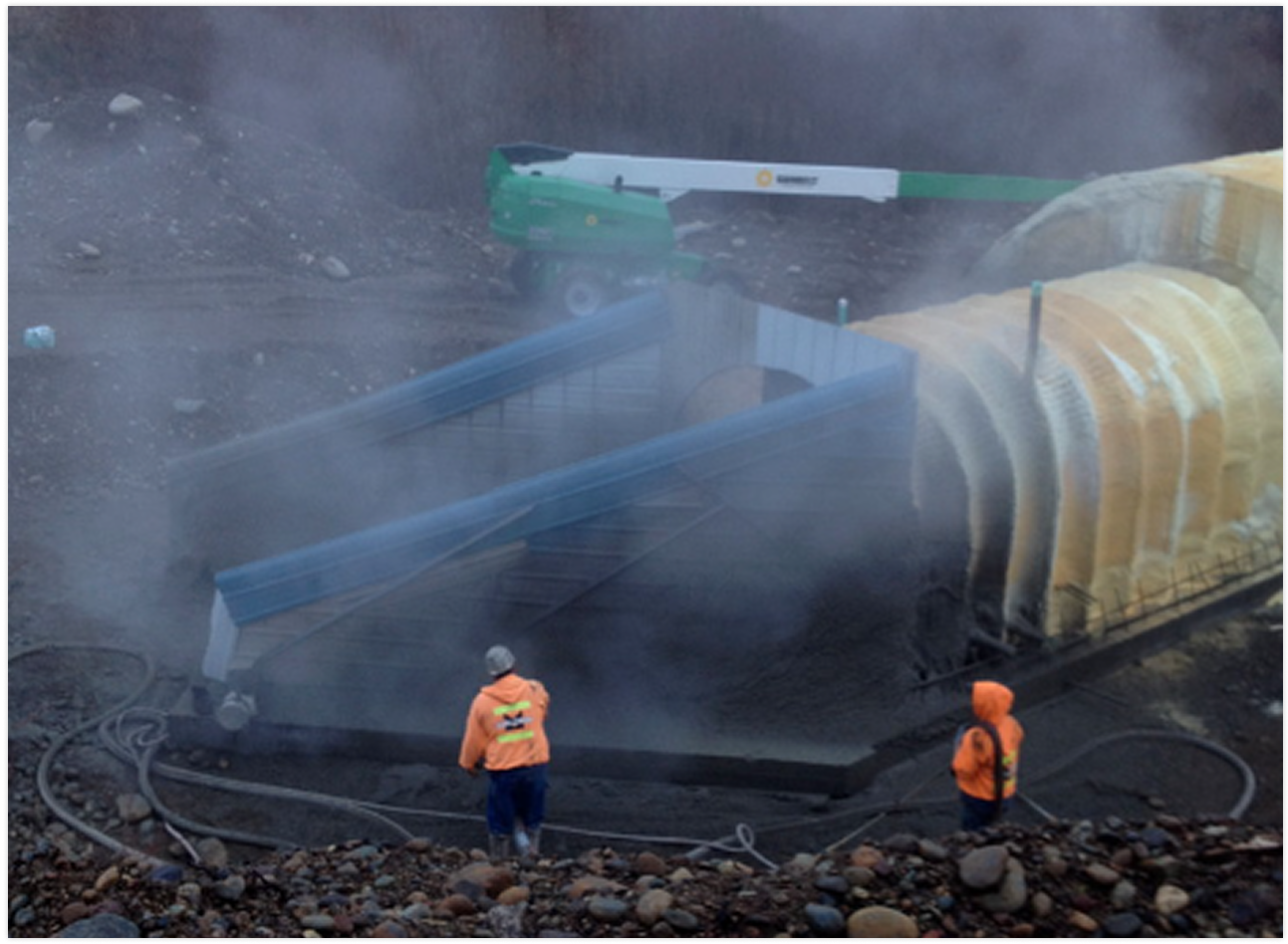 Construction site with two workers in orange safety jackets and helmets, large metallic structure, and equipment, with dust and dirt surrounding the area.