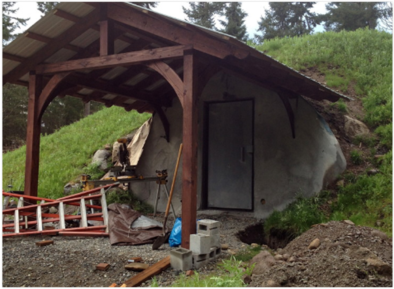 Construction of a small building with a wooden and metal roof, built into a hillside with a concrete or stone wall, surrounded by construction tools and materials.