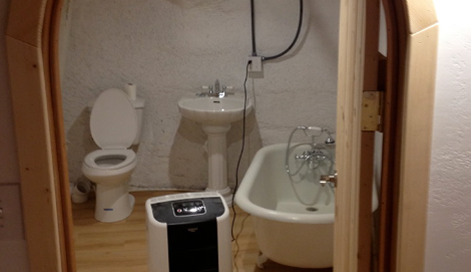A small bathroom with a toilet, a pedestal sink, a clawfoot bathtub, and a portable air purifier. The bathroom has wooden flooring and a textured white wall.