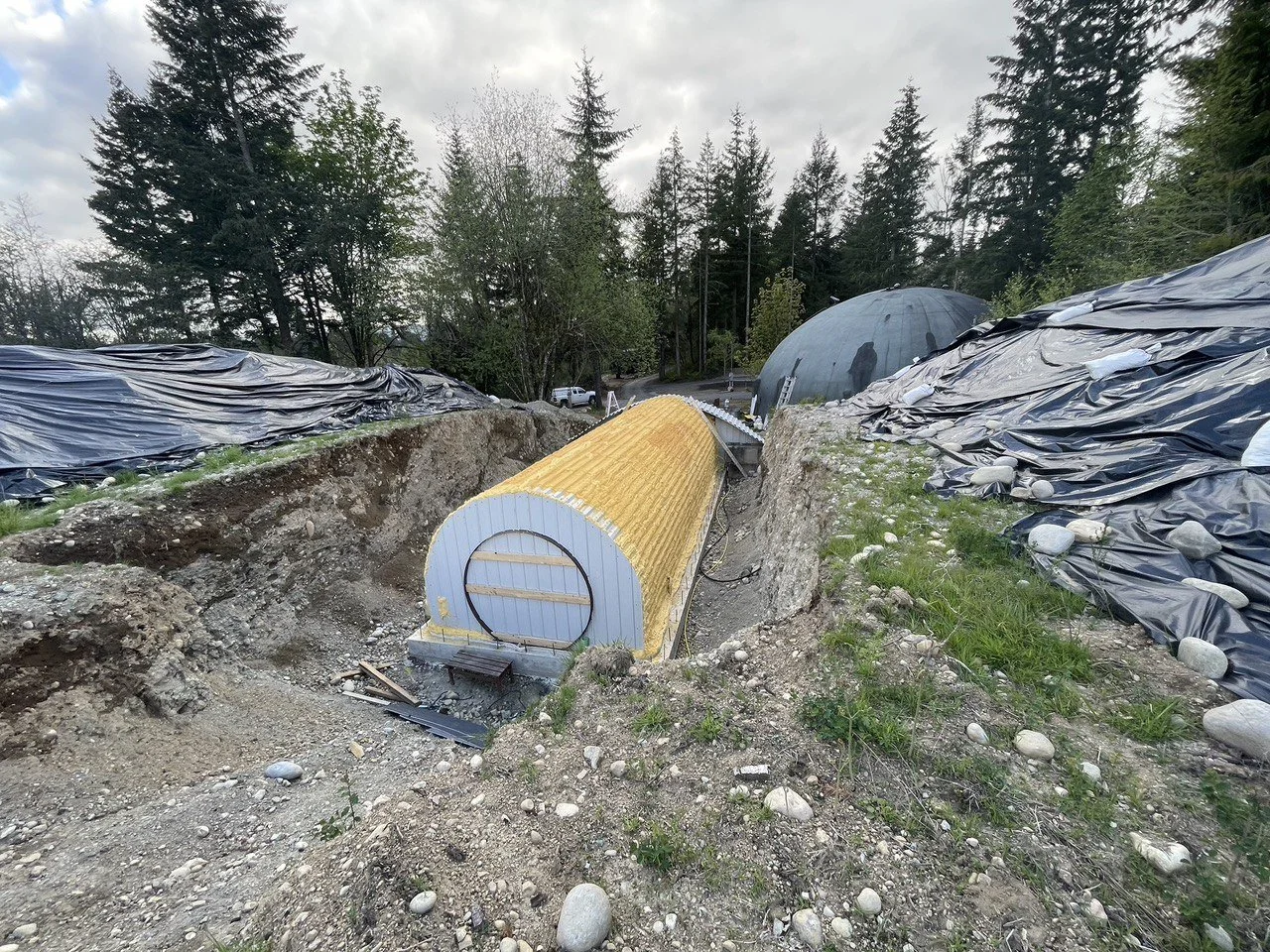 Construction site with an underground tunnel structure in a trench during daytime, surrounded by rocky and grassy terrain with trees and cloudy sky in the background.