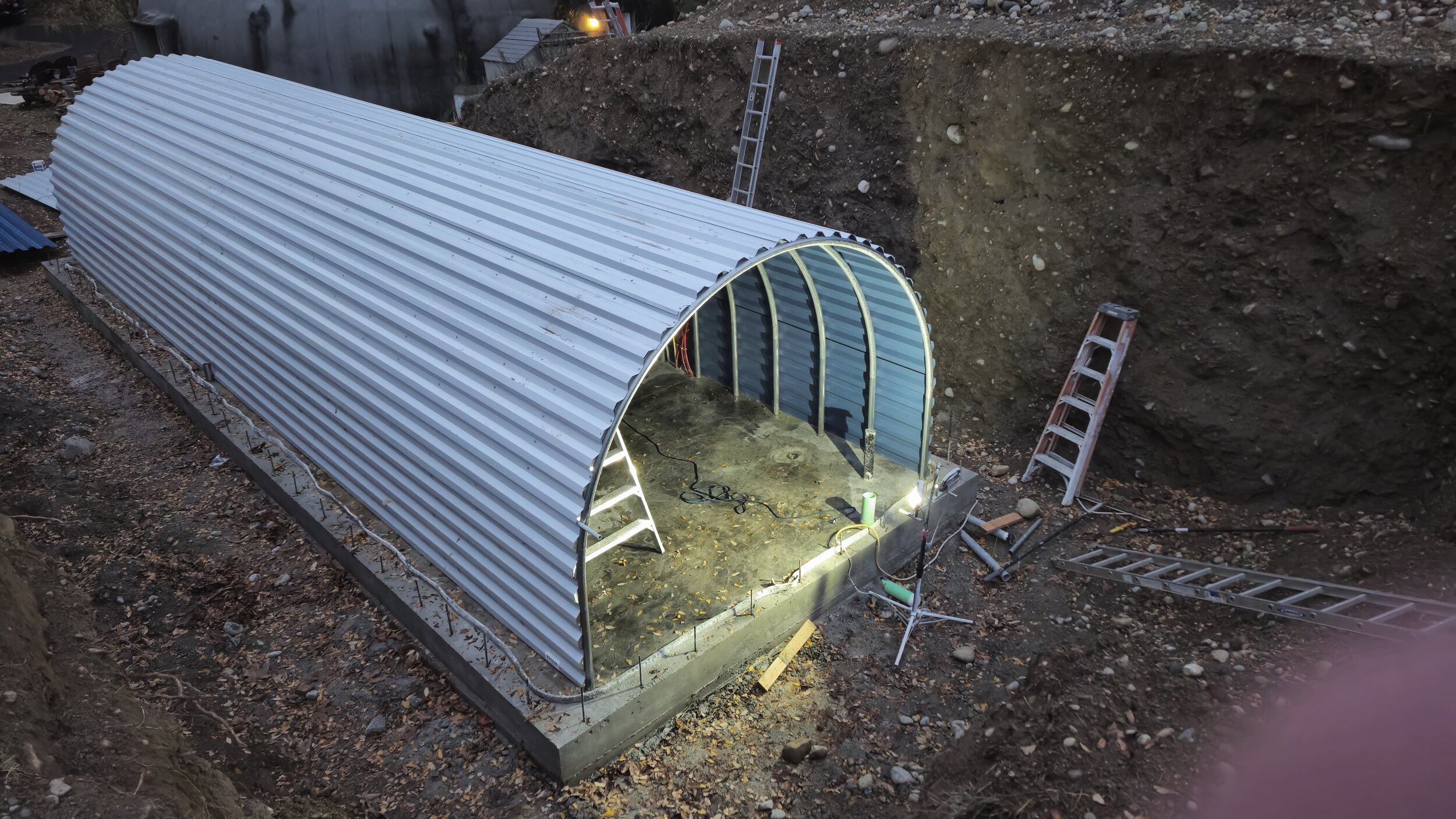 Construction site with a metal-framed, arched structure being built, surrounded by ladders and construction tools, with the ground cleared and excavated around it.