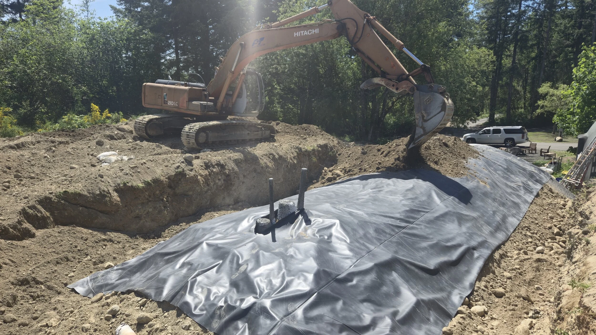 A construction site with a Hitachi excavator leveling earth over a plastic drainage or waterproofing sheet on a slope. There are trees and a parked vehicle in the background.