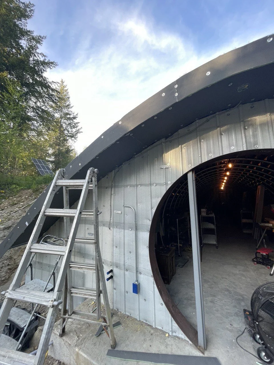 Construction of a cylindrical building with metal walls and a large, curved opening, surrounded by trees and blue sky, with a ladder and construction tools nearby.