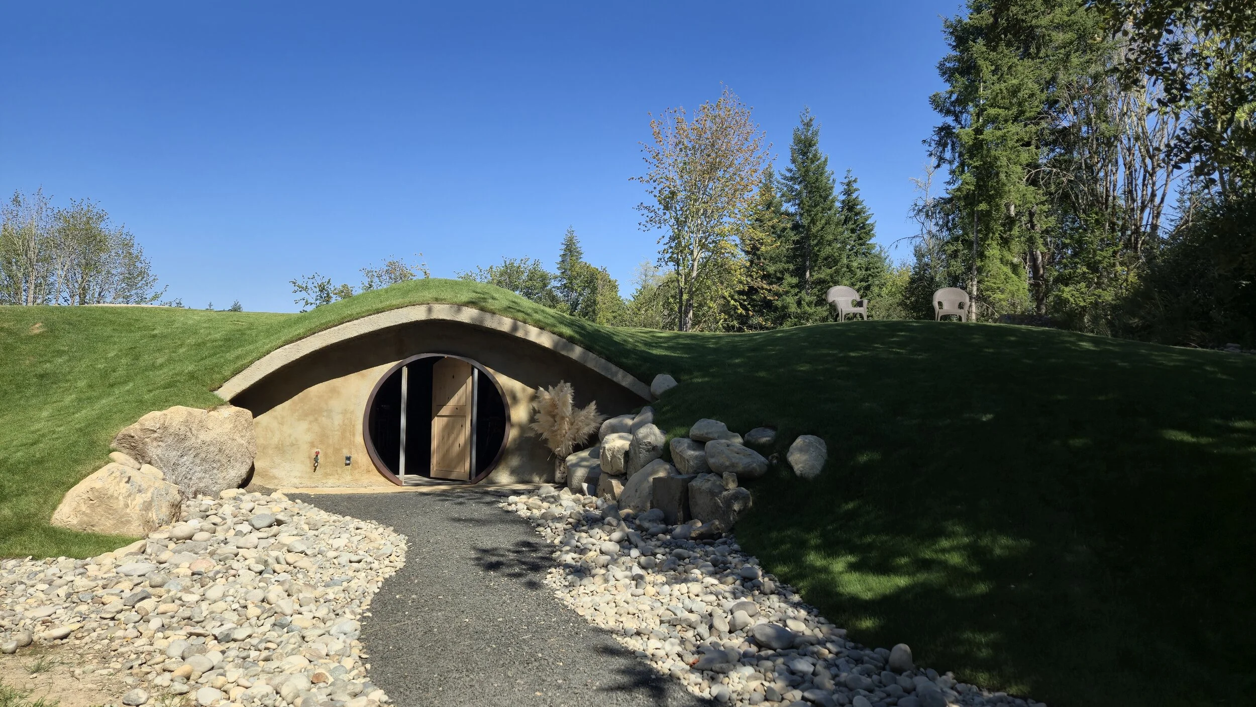 A moss-covered hill with a circular, earth-colored entrance and a gravel pathway leading to it, set in a lush green landscape with trees and two lawn chairs on the hilltop, under a clear blue sky.