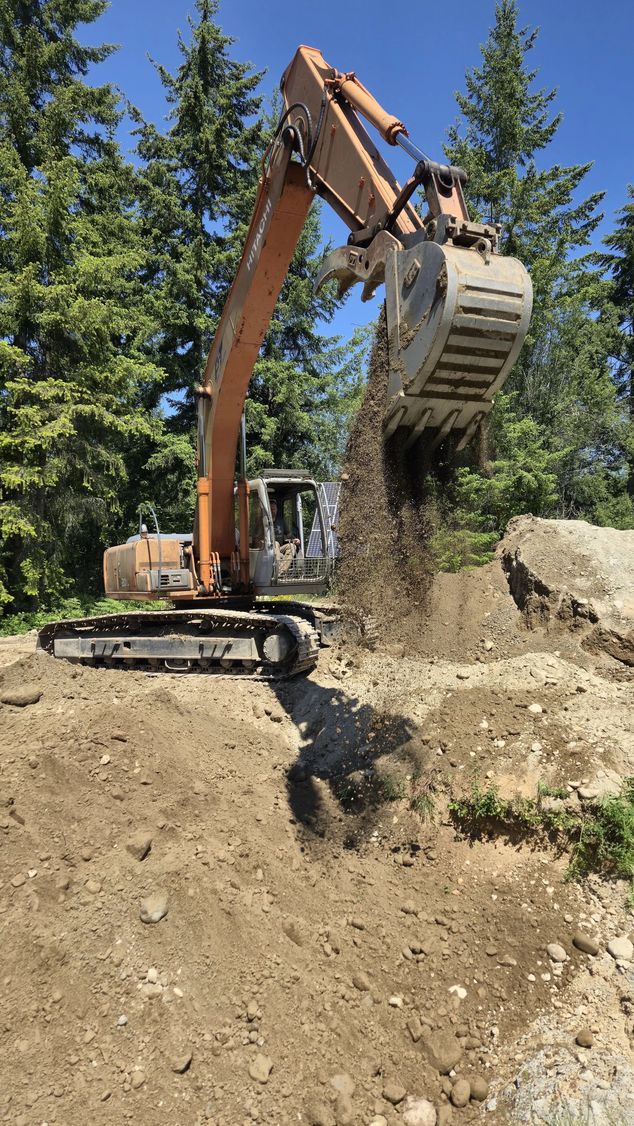 A large construction excavator digging into the dirt on a construction site with trees and a blue sky in the background.