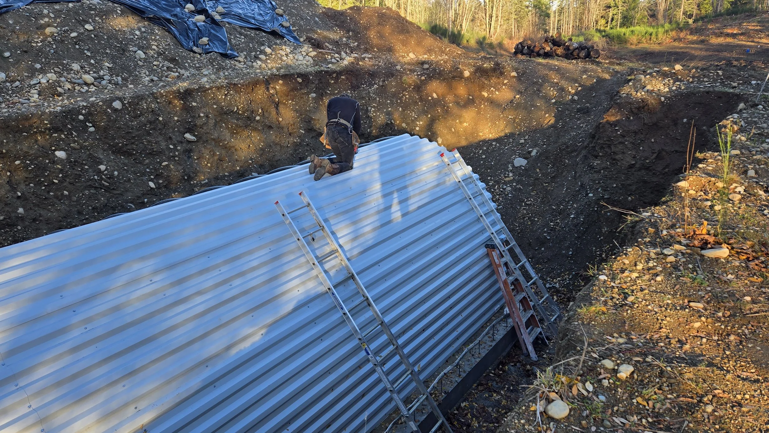 A construction worker installing a corrugated metal pipe in a large trench during daylight hours. The worker is kneeling on top of the pipe, which is supported by ladders, with a forested background.