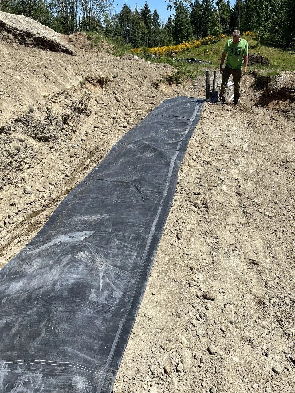 A man working outdoors on a construction site, laying black plastic sheeting on a dirt pathway surrounded by soil and trees, with flowers in the background.