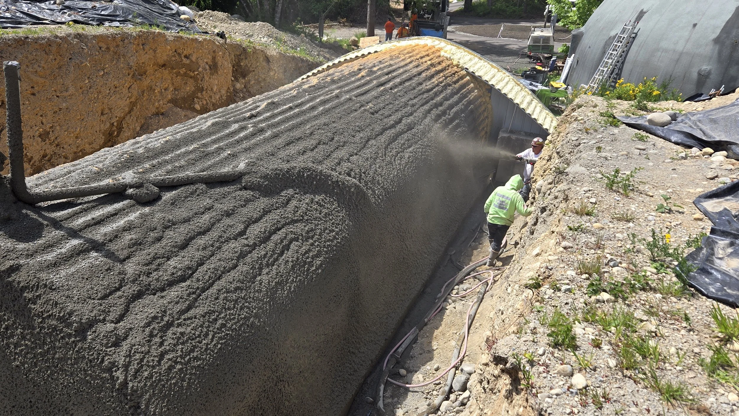 Workers paving a large concrete pipeline in an outdoor construction site, with trees and equipment in the background.
