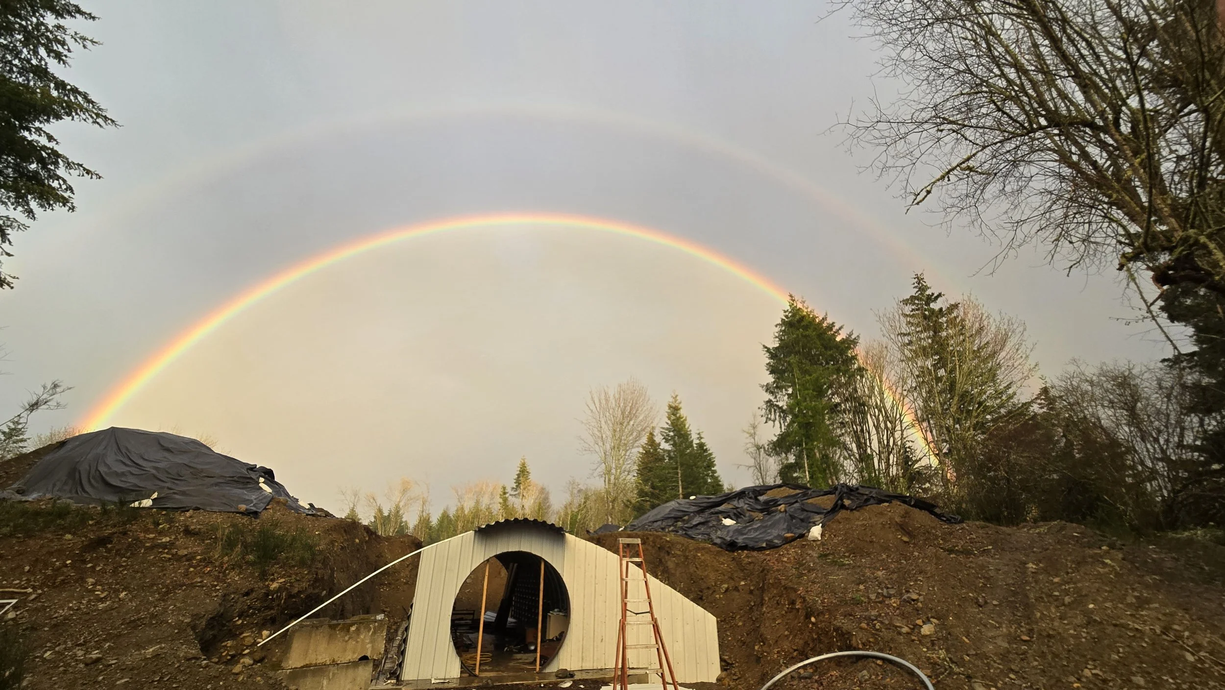 Construction site with a rainbow in the sky, trees in the background, and a partially built tunnel entrance in the foreground.