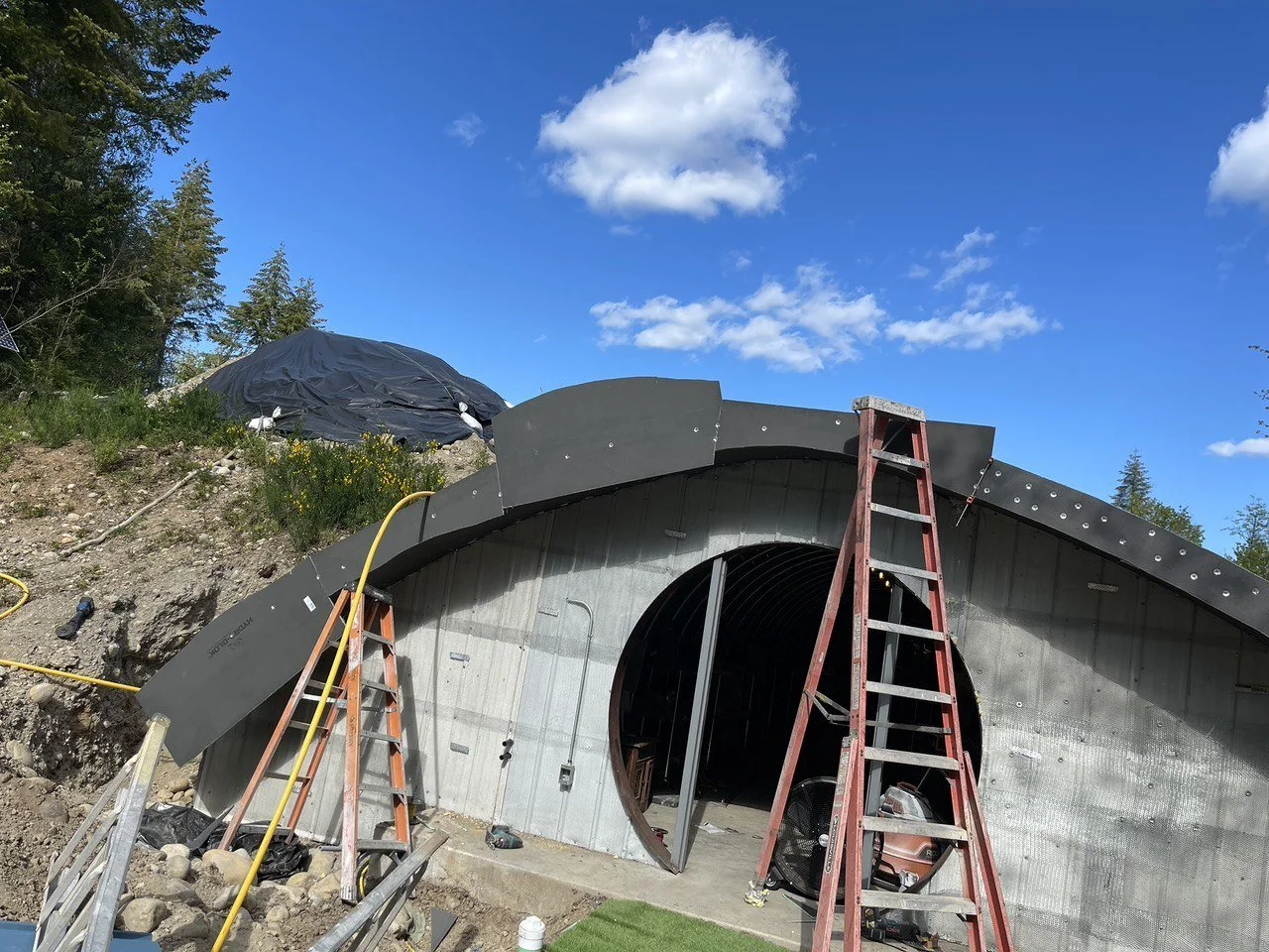 Construction site of a tunnel through a hillside, with ladders and construction materials in front, and a partially built dark metal arch-shaped structure.