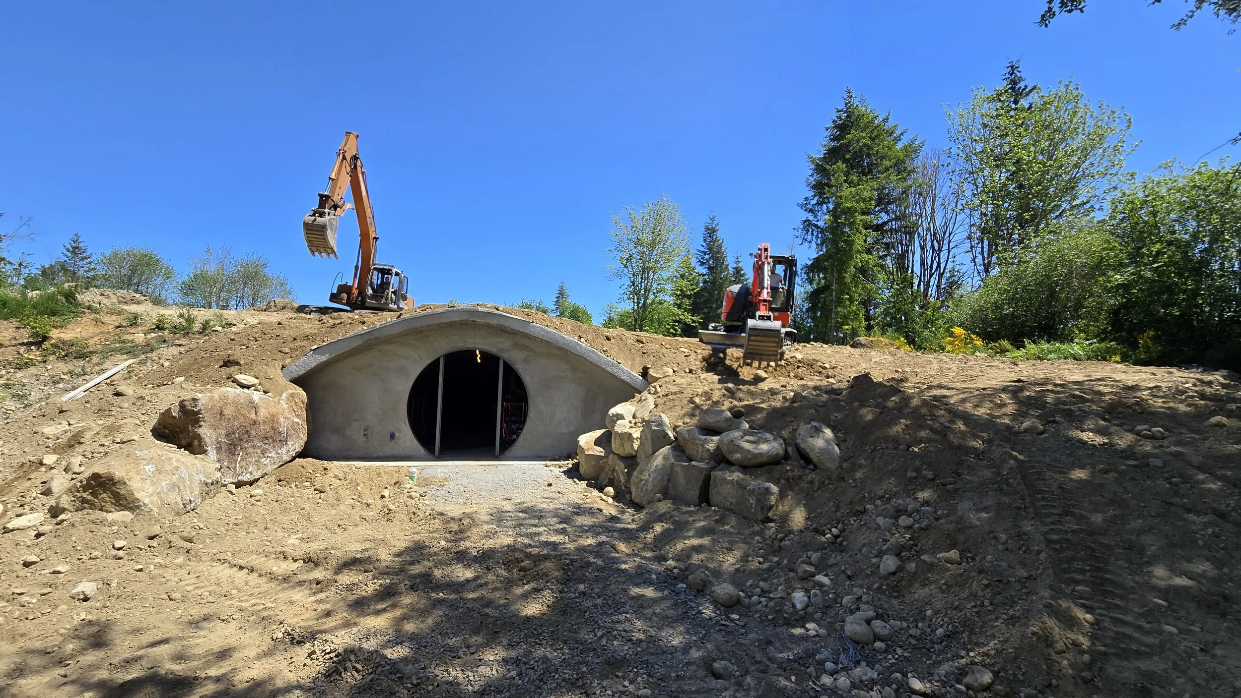 Construction site with two excavators on dirt above a concrete tunnel entrance, surrounded by rocks, trees, and a clear blue sky.