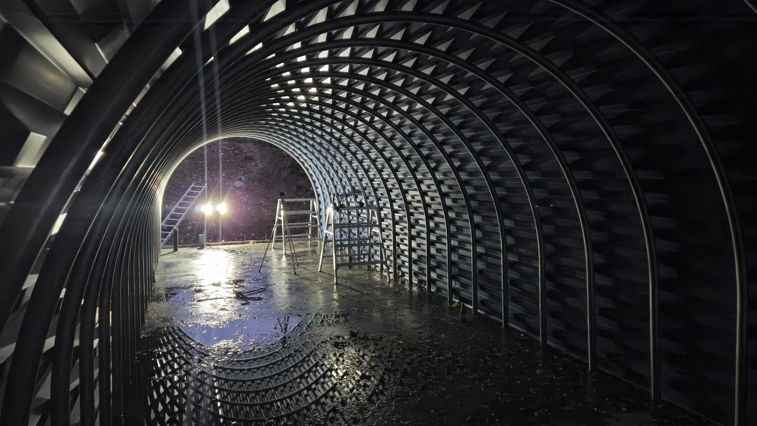 Inside a construction tunnel with curved metal framework, construction tools like ladders and scaffolding are present, and there are water puddles on the ground reflecting light, indicating ongoing construction work.