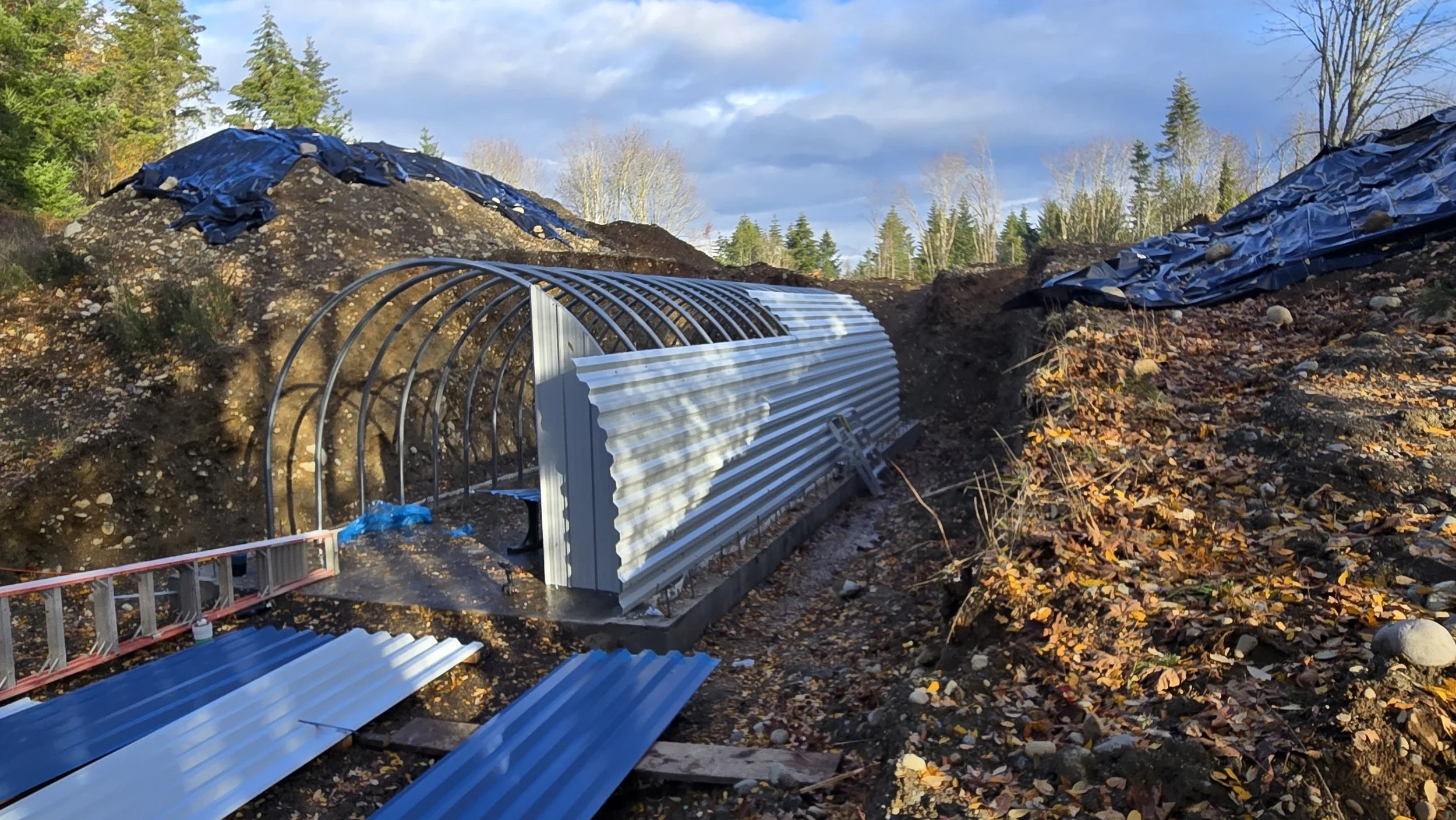 A construction site in a wooded area showing a partially built metal structure, with black plastic covers on mounds of dirt, and surrounded by autumn leaves and trees.
