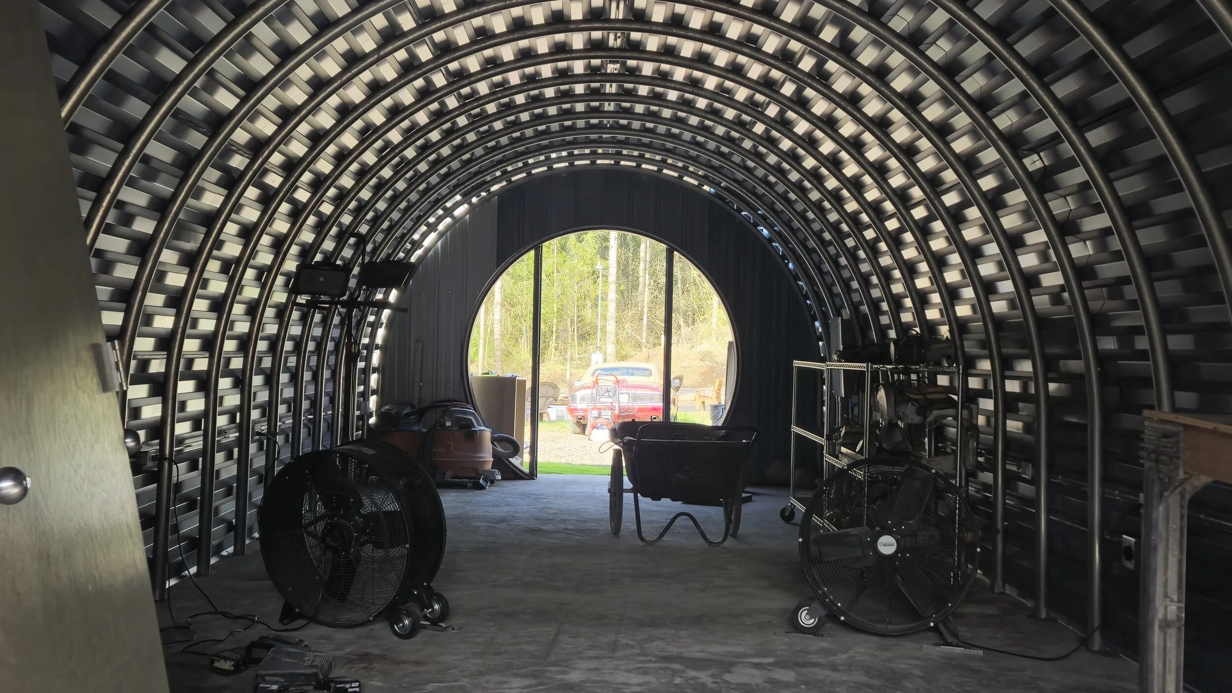 View inside a metal tunnel structure with tools, fans, and equipment, leading to an open door with a red truck and trees outside