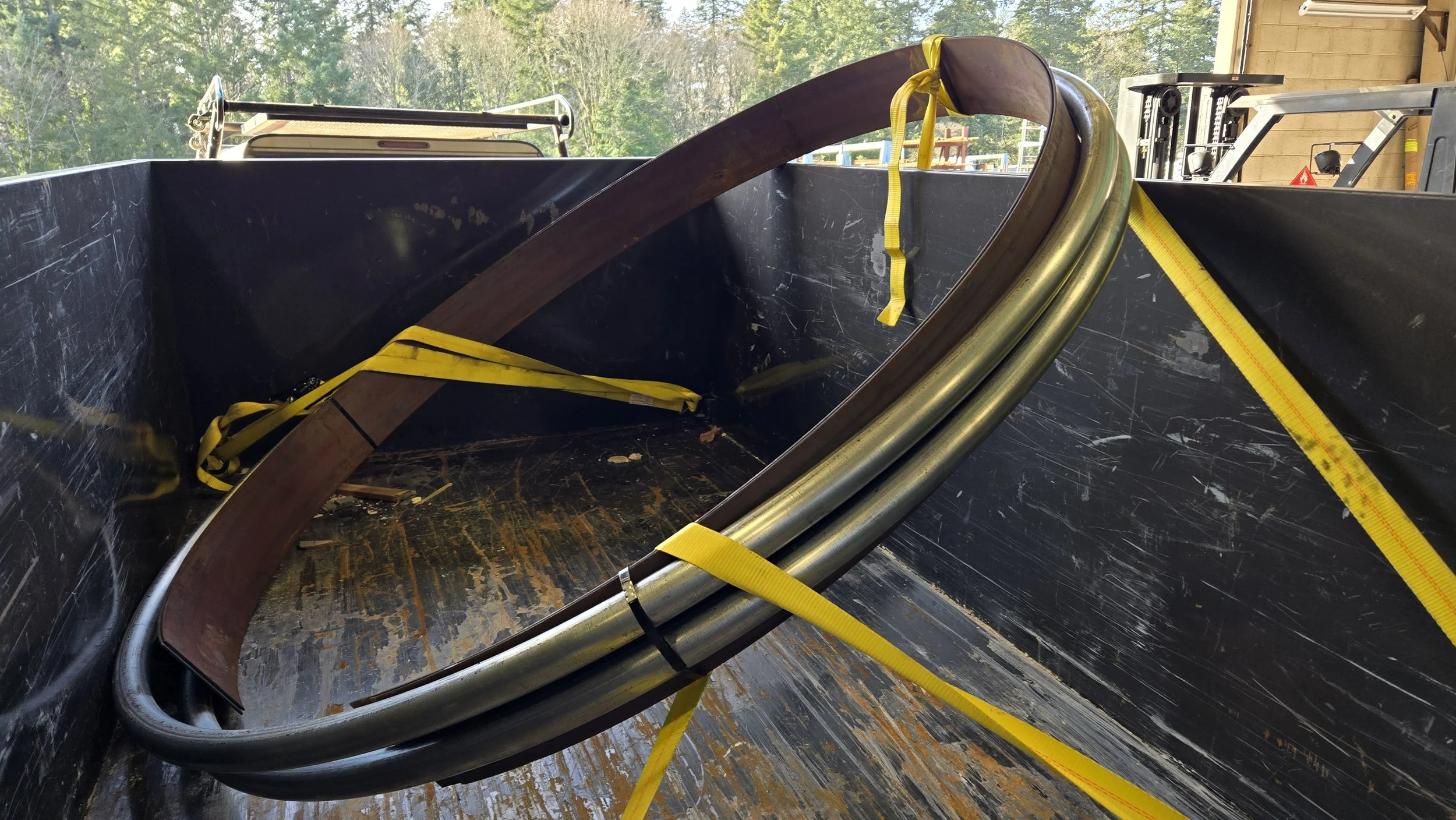 Steel pipes and a curved metal part secured with yellow straps inside a black industrial container, outdoor scene with trees in the background.