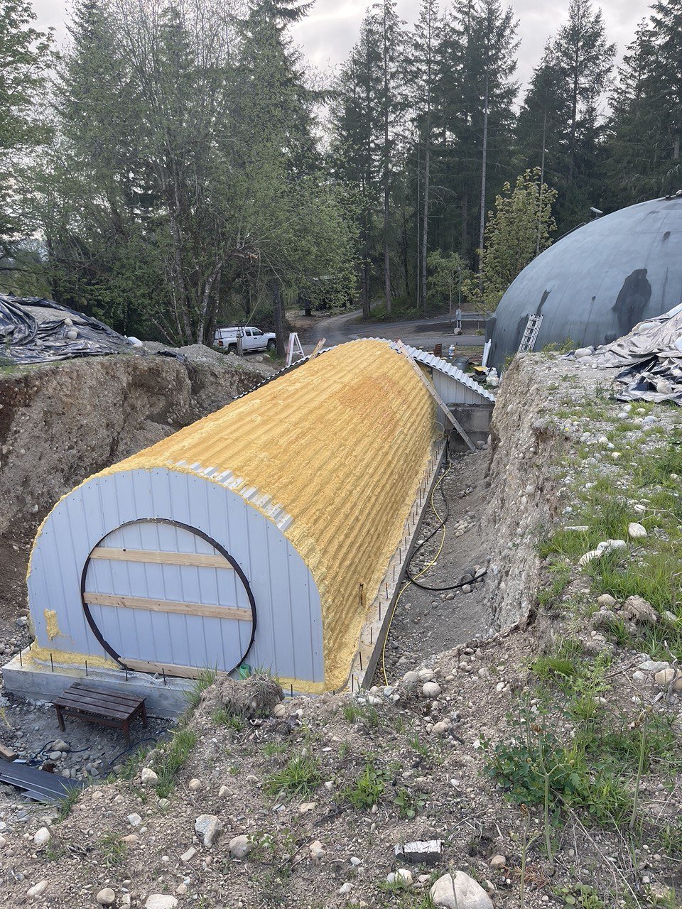 Construction of a large, tunnel-shaped building with a ribbed, yellow insulation material on the exterior, surrounded by dirt and rocky terrain.