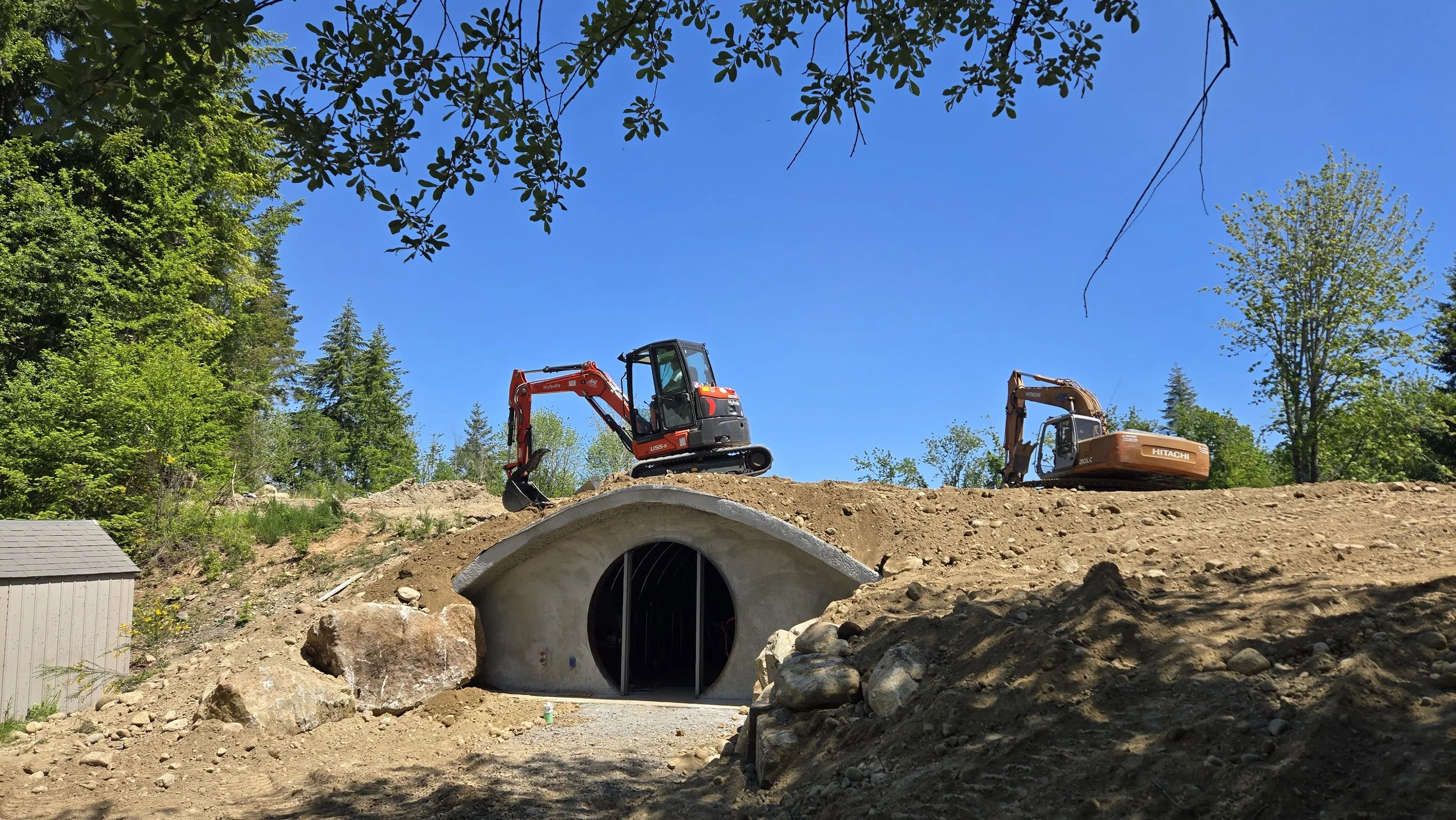 Construction site with two excavators on a dirt hill, one red and one yellow, near a concrete tunnel entrance surrounded by rocks and trees under a blue sky.
