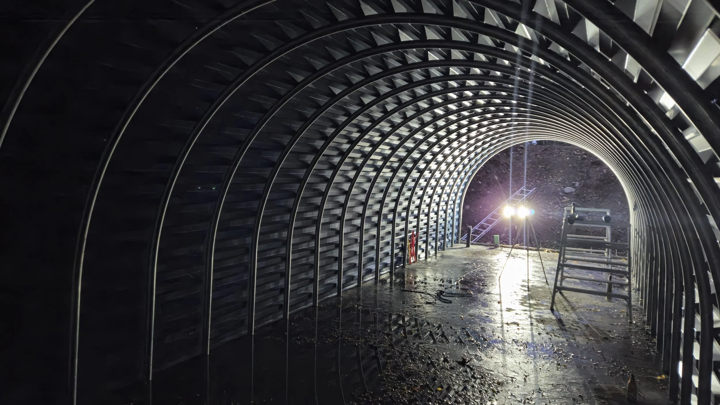 Inside a metal tunnel under construction with bright light at the far end, a ladder, and construction equipment.