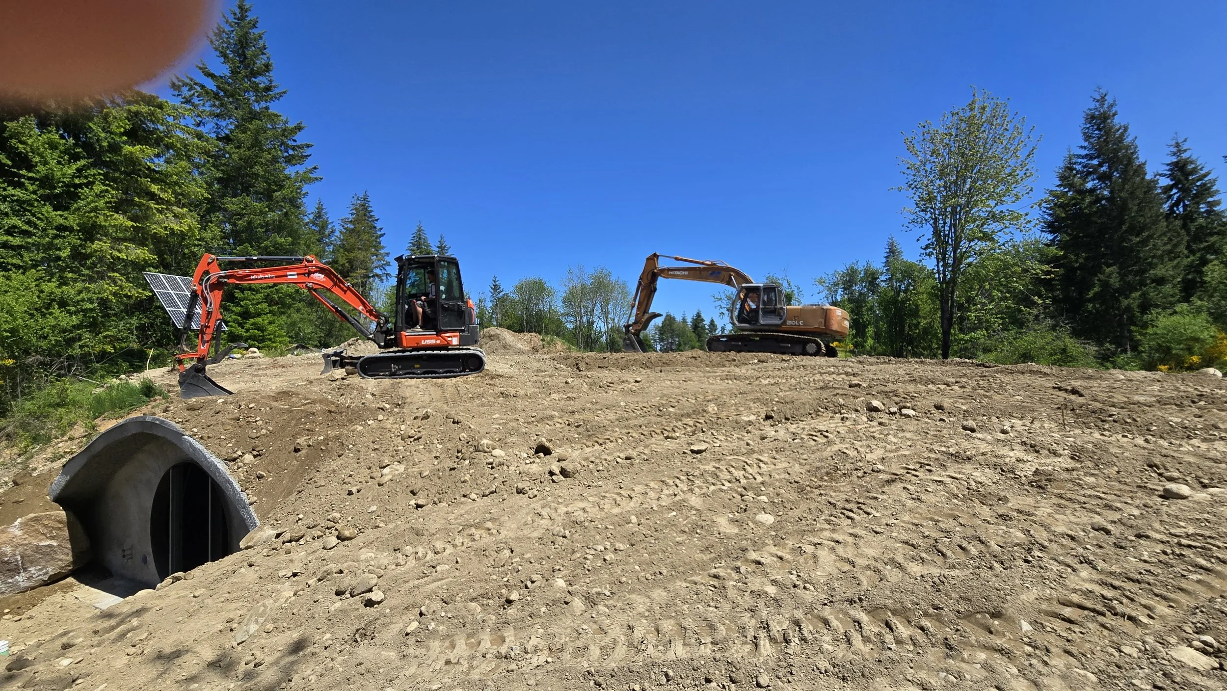 Two excavators work on a construction site amidst a backdrop of trees and a clear blue sky.
