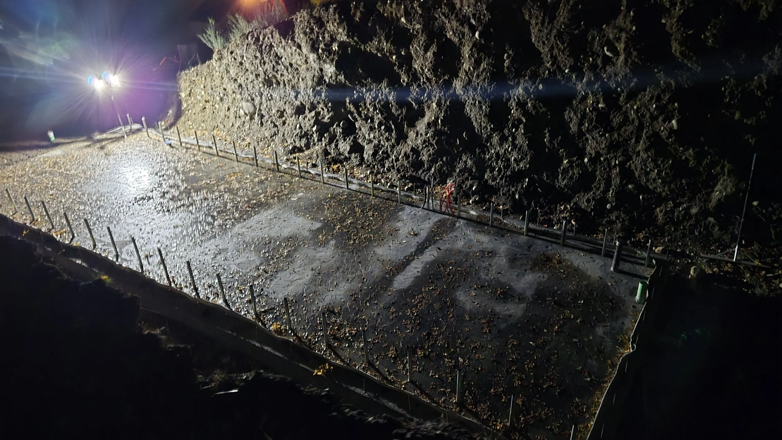 Night view of a construction site with a concrete foundation and steel reinforcements, illuminated by bright lights, with a dirt mound and trees in the background.