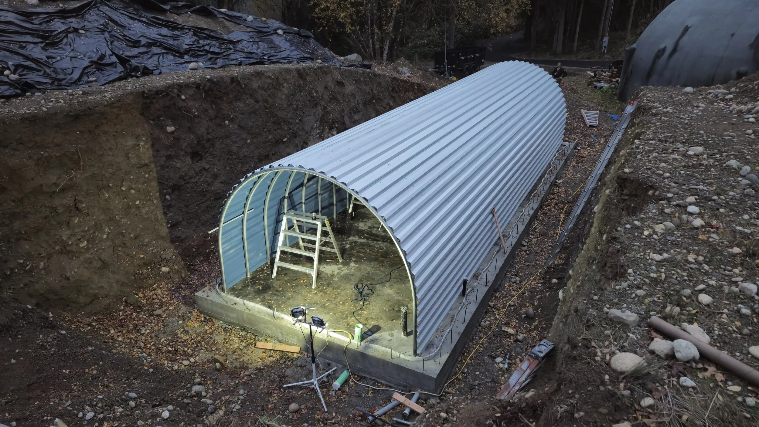 Under construction, a tunnel-shaped greenhouse made of metal and plastic panels in a dugout area, with construction tools and ladder inside.