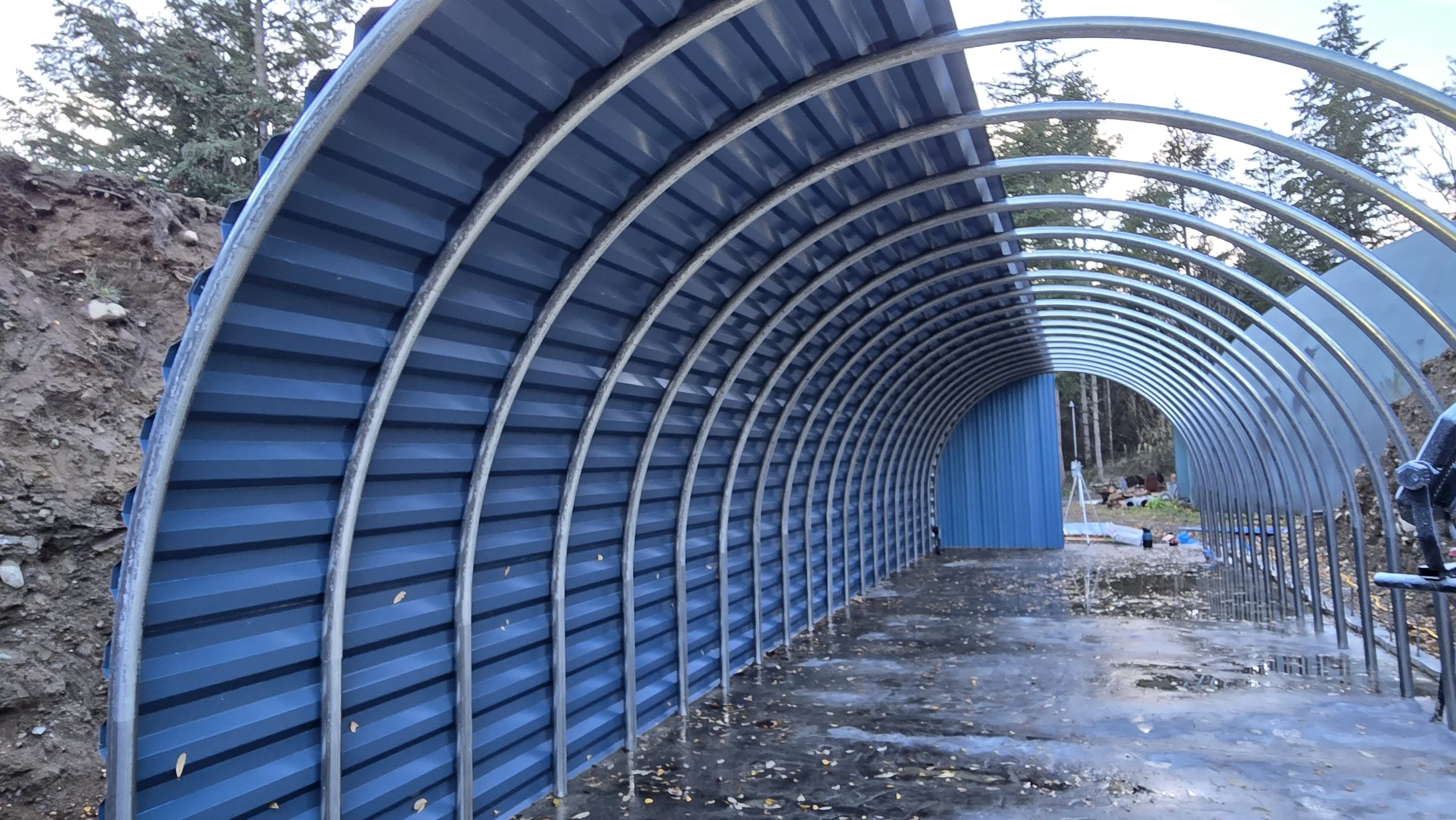 A blue metal tunnel under construction with a curved arch structure, on a muddy ground with a forest in the background.